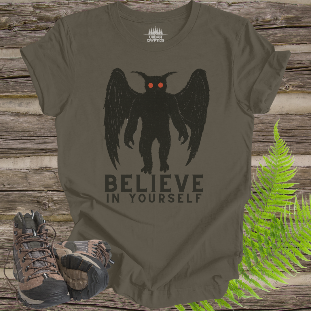 T-shirt with a creature graphic and 'Believe in Yourself' text on a wooden background with boots and ferns.
