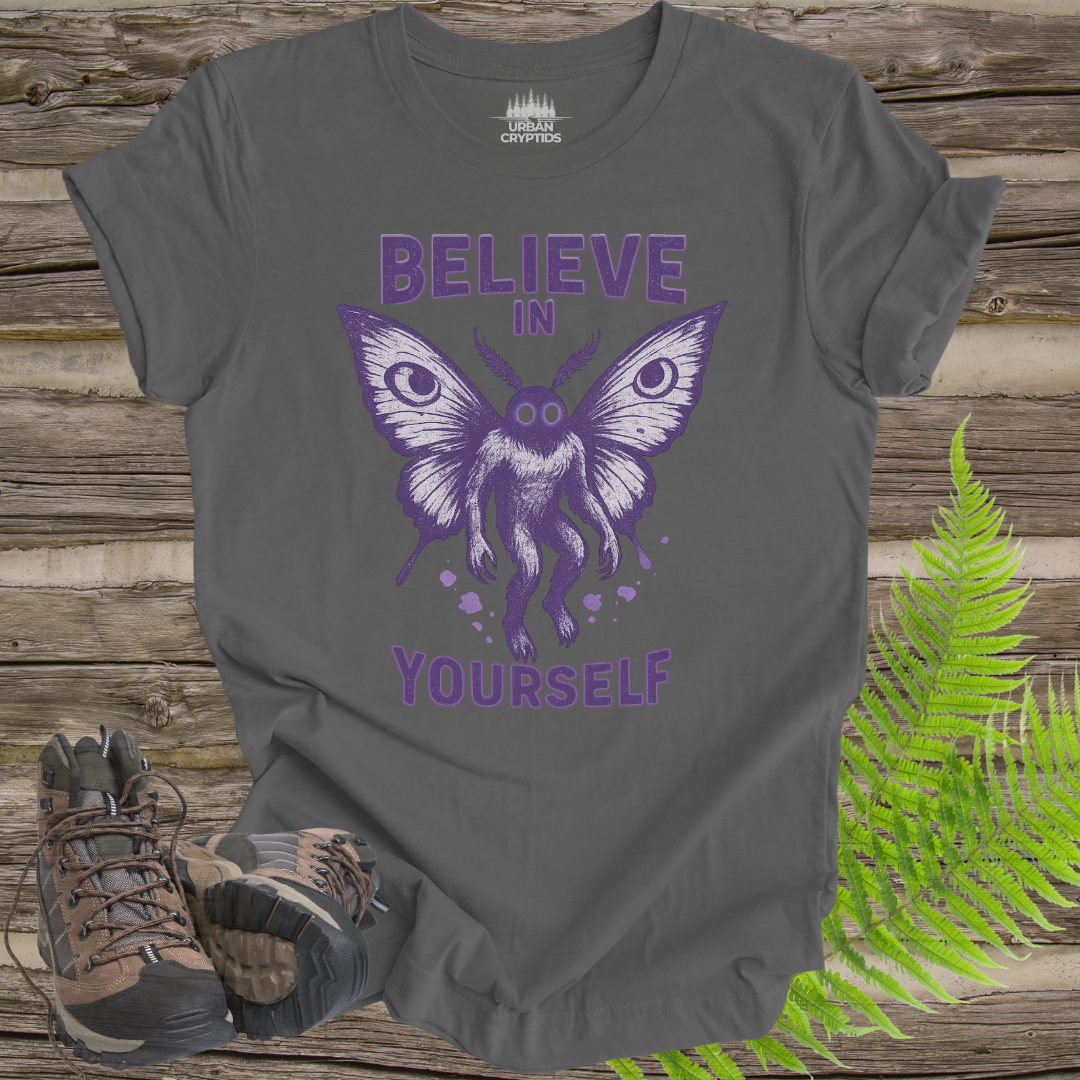 Gray t-shirt with purple butterfly graphic and 'Believe in Yourself' text on a wooden surface with hiking boots and ferns.