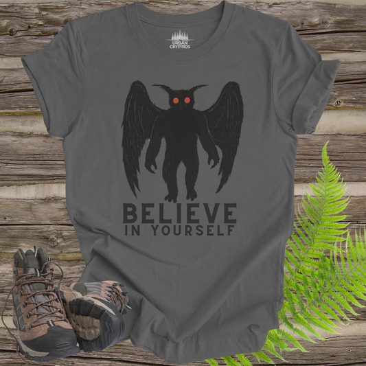 Gray t-shirt with a black creature graphic and 'BELIEVE IN YOURSELF' text on a wooden surface with hiking boots and ferns.