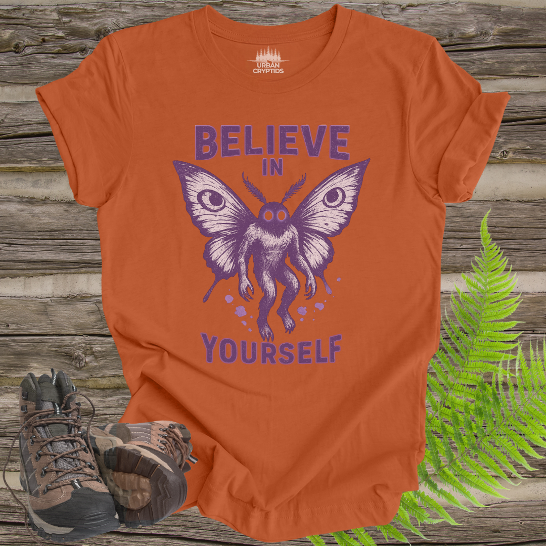 Orange t-shirt with purple butterfly graphic and 'Believe in Yourself' text on a wooden surface with hiking boots and ferns.
