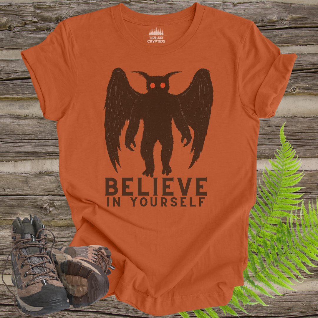 Orange t-shirt with a black owl graphic and 'BELIEVE IN YOURSELF' text on a wooden surface with hiking boots and ferns.