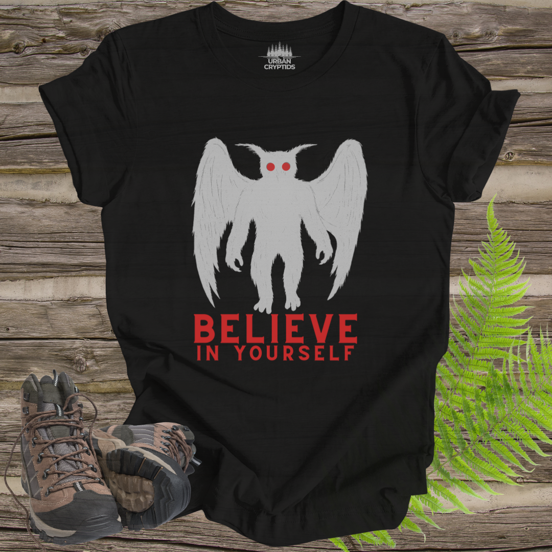 Black t-shirt with a white creature graphic and 'BELIEVE IN YOURSELF' text on a wooden surface with hiking boots and ferns.