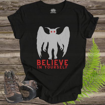 Black t-shirt with a white creature graphic and 'BELIEVE IN YOURSELF' text on a wooden surface with hiking boots and ferns.