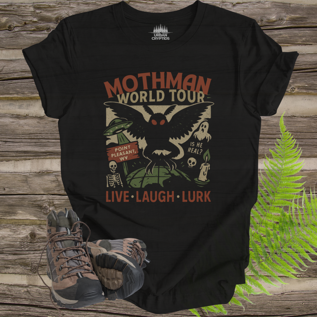 Black t-shirt with Mothman graphic design on a wooden surface with hiking boots and ferns.