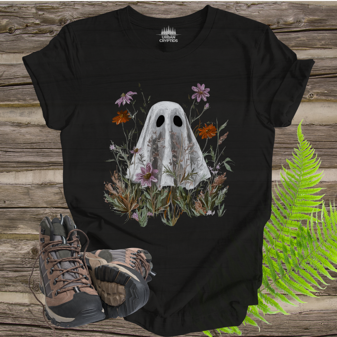 Black t-shirt with ghost and floral design on a wooden surface with hiking boots and ferns.