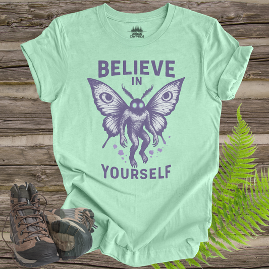 Mint green t-shirt with butterfly graphic and 'Believe in Yourself' text on a wooden surface with hiking boots and fern.