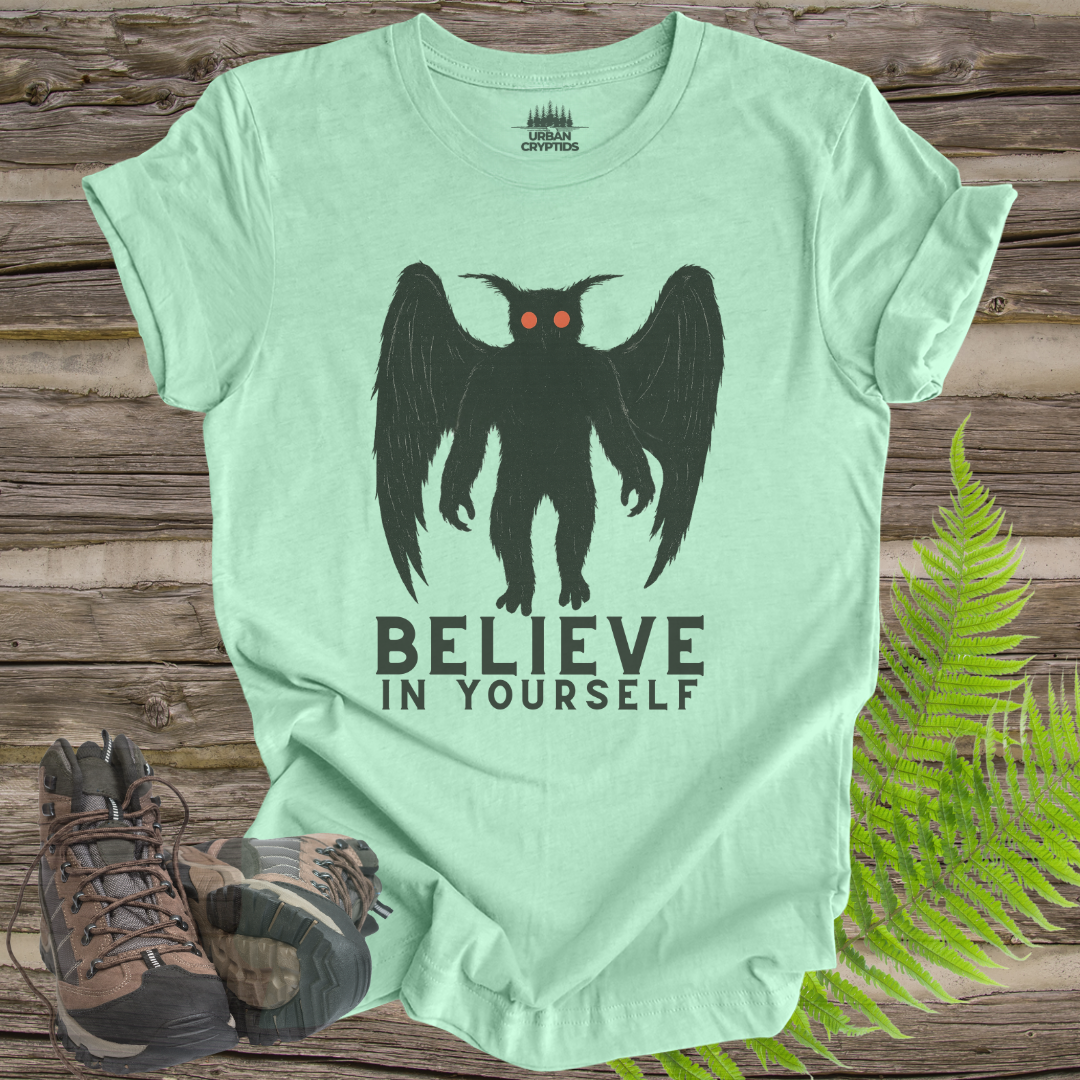 Mint green t-shirt with a black creature graphic and 'Believe in Yourself' text on a wooden surface with hiking boots and ferns.