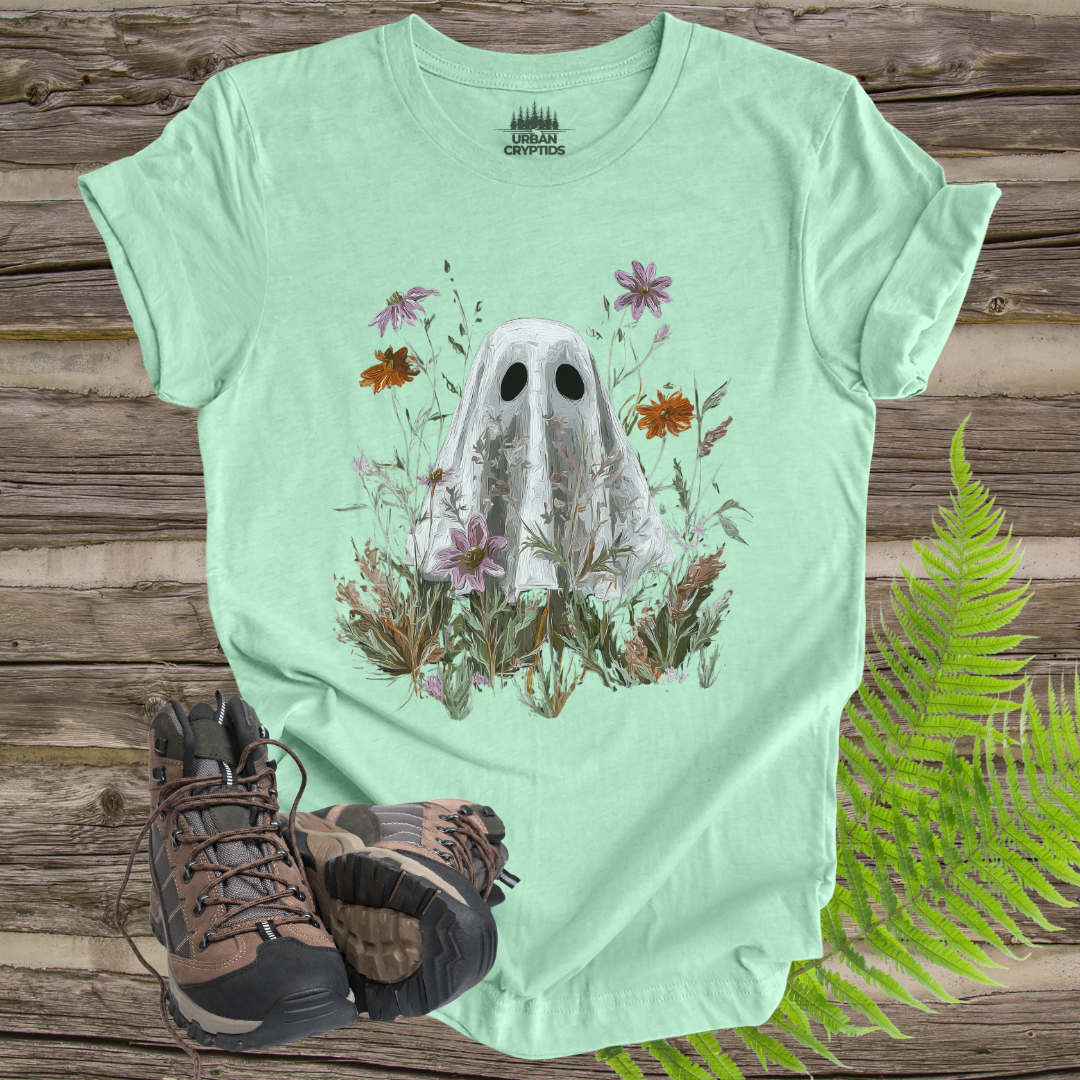 Light green t-shirt with ghost and floral design on a wooden surface with hiking boots and ferns.