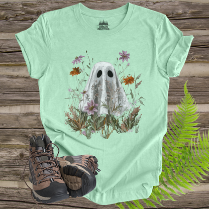 Light green t-shirt with ghost and floral design on a wooden surface with hiking boots and ferns.