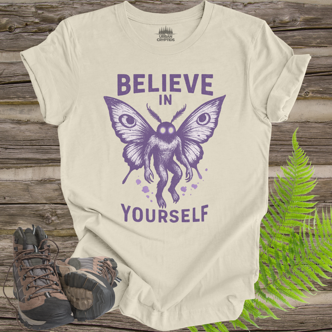 Beige t-shirt with purple butterfly graphic and 'Believe in Yourself' text on a wooden surface with hiking boots and ferns.