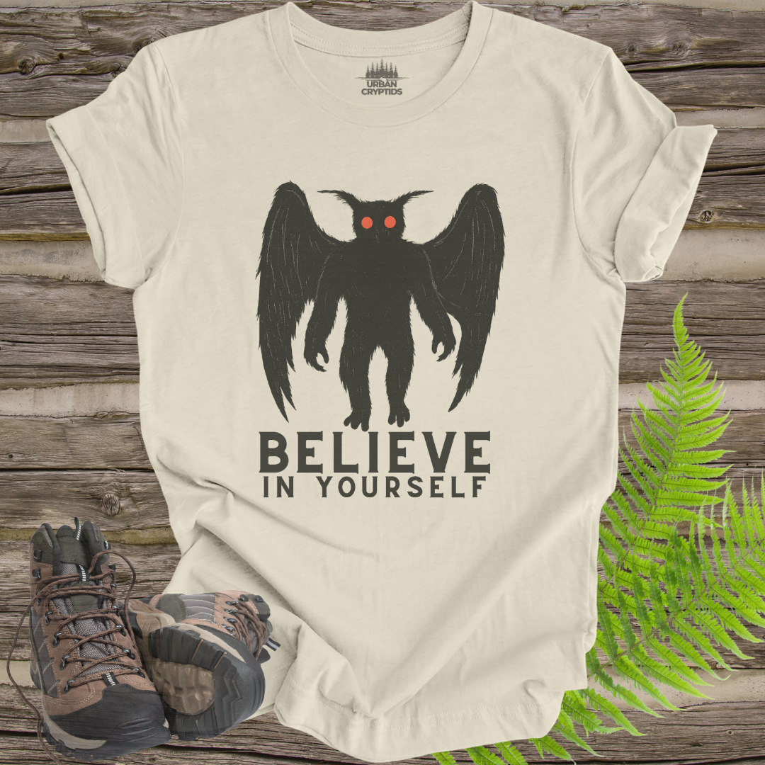 Beige t-shirt with a black creature graphic and 'BELIEVE IN YOURSELF' text on a wooden surface with hiking boots and ferns.
