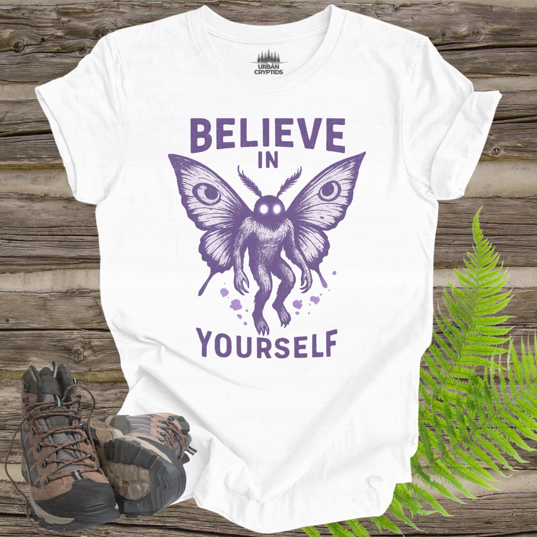 White t-shirt with purple butterfly graphic and 'Believe in Yourself' text on a wooden surface with hiking boots and fern.