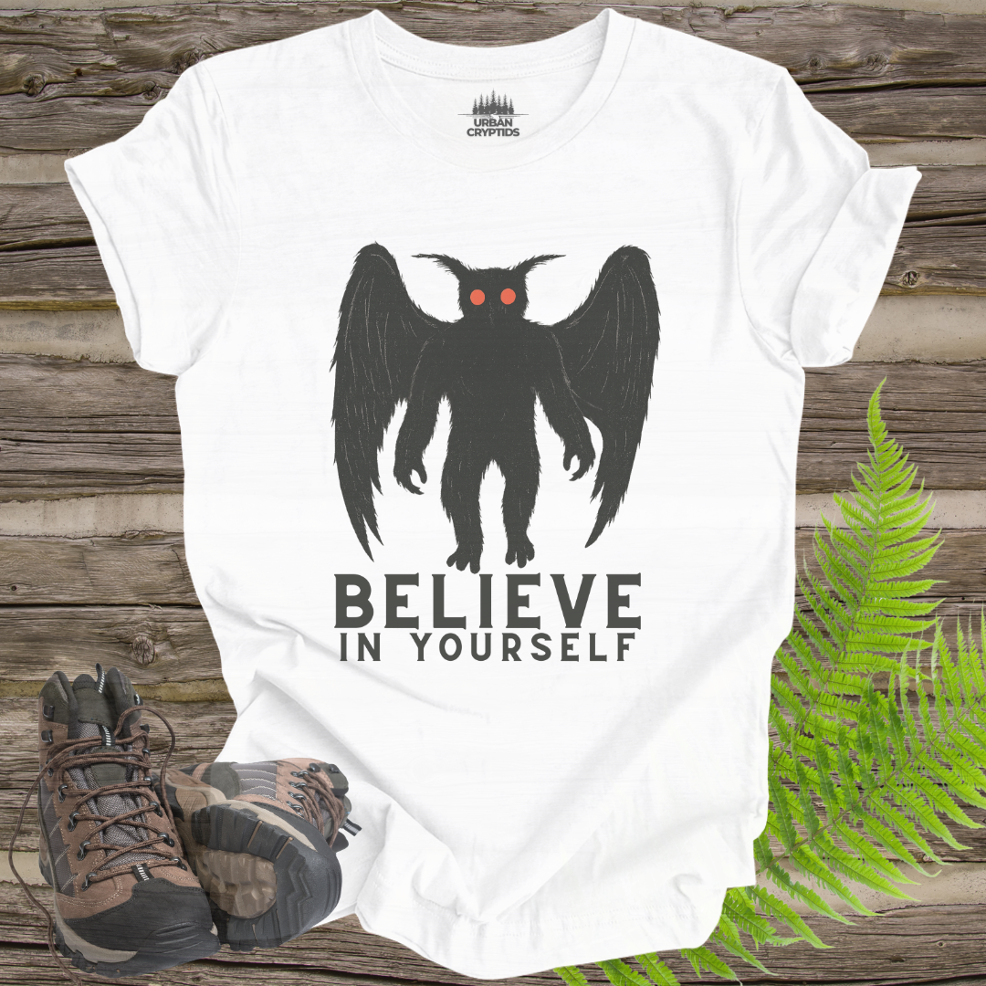 White t-shirt with a black creature graphic and 'BELIEVE IN YOURSELF' text on a wooden surface with hiking boots and ferns.