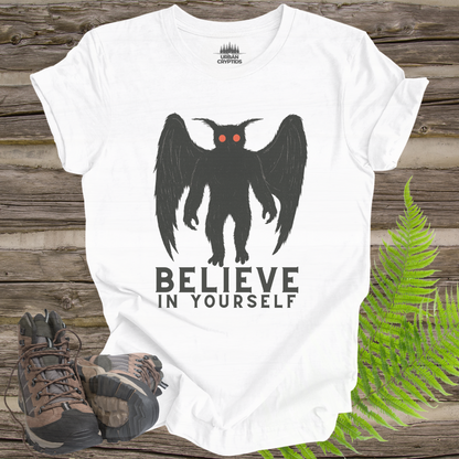 White t-shirt with a black creature graphic and 'BELIEVE IN YOURSELF' text on a wooden surface with hiking boots and ferns.