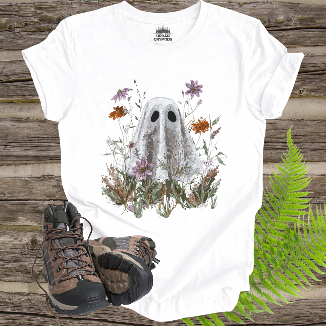 White t-shirt with ghost and floral design on a wooden surface with hiking boots and ferns.